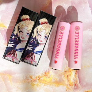 May include: Two black and white cardboard boxes with a cartoon image of a sailor girl holding a black cat. The boxes are labeled "Vinnabelle". Two pink lipstick tubes with gold caps are labeled "Vinnabelle" with a pink heart and a star.