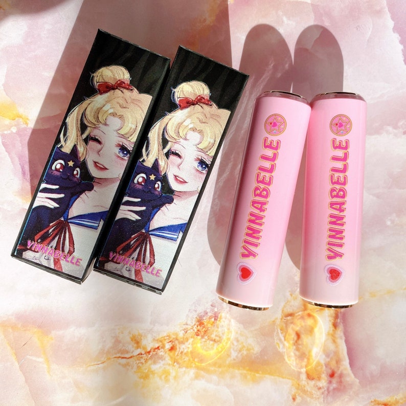May include: Two black and white cardboard boxes with a cartoon image of a sailor girl holding a black cat. The boxes are labeled "Vinnabelle". Two pink lipstick tubes with gold caps are labeled "Vinnabelle" with a pink heart and a star.