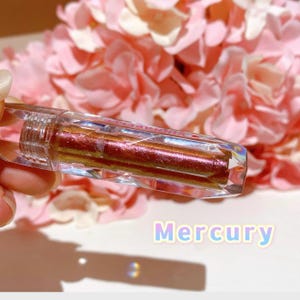 May include: A clear, faceted, tube-shaped container with a metallic, iridescent, red-gold liquid inside. The container is labeled "Mercury".