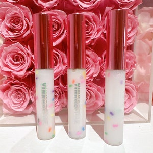 May include: Three clear lip gloss tubes with a white base and a pink lid. The glosses have colorful sprinkles inside. The tubes are sitting on a white surface in front of a bouquet of pink roses.