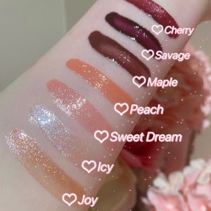 May include: A close-up of eight different lip glosses swatched on a person's arm. The lip glosses are labeled with the names "Joy", "Icy", "Sweet Dream", "Peach", "Maple", "Savage", and "Cherry".