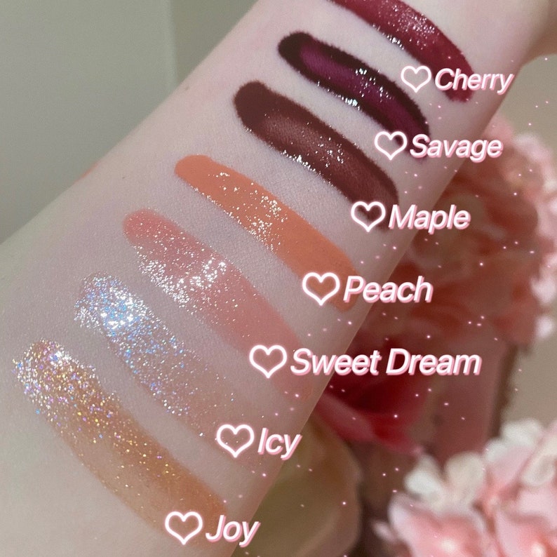 May include: A close-up of eight different lip glosses swatched on a person's arm. The lip glosses are labeled with the names "Joy", "Icy", "Sweet Dream", "Peach", "Maple", "Savage", and "Cherry".