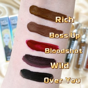 May include: Swatches of five liquid lipsticks in shades of brown, red, and black, labeled "Rich", "Boss Up", "Bloodshot", "Wild", and "Over You". The lipsticks are applied to the skin, showcasing their colors. The image also includes the lipstick tubes.
