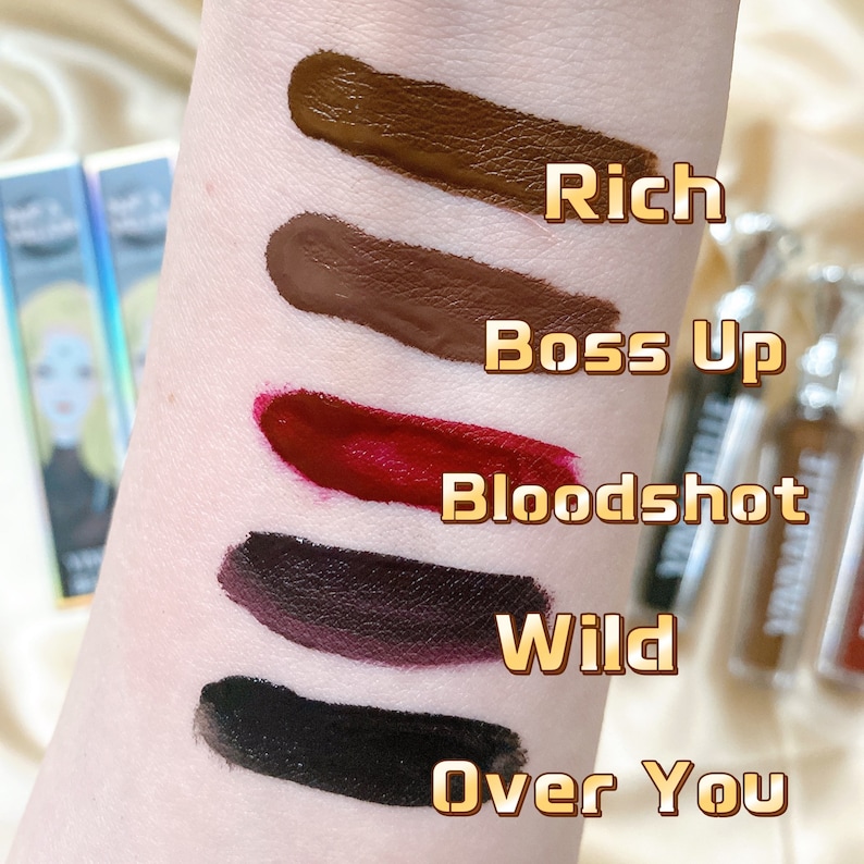 May include: Swatches of five liquid lipsticks in shades of brown, red, and black, labeled "Rich", "Boss Up", "Bloodshot", "Wild", and "Over You". The lipsticks are applied to the skin, showcasing their colors. The image also includes the lipstick tubes.
