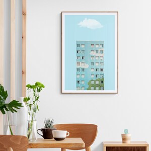 May include: A framed art print depicting a light blue apartment building with numerous windows, set against a pale blue sky with a white cloud. The print is in a brown frame, displayed in a room with wooden furniture and greenery.