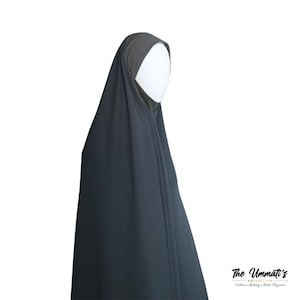 Könnte beinhalten: Ein dunkelgrauer Hidschab, eine traditionelle islamische Kopfbedeckung, ist über einem weißen Schaufensterkopf drapiert. Der Hidschab hat eine kontrastierende graue Kopfbedeckung und ein fließendes Design. Das Logo "The Ummatī's" ist unten rechts sichtbar.