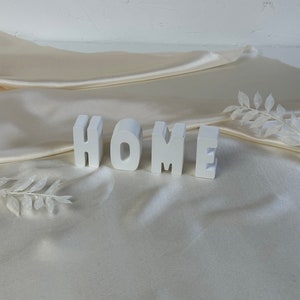 Könnte beinhalten: Weiße Holzbuchstaben mit der Aufschrift "HOME" liegen auf einem cremefarbenen Satin-Stoff. Die Buchstaben im Blockstil stehen aufrecht. Dekorative weiße Zweige sind auf dem Stoff platziert und verleihen der Komposition Eleganz. Der Hintergrund ist ein weiches Off-White.