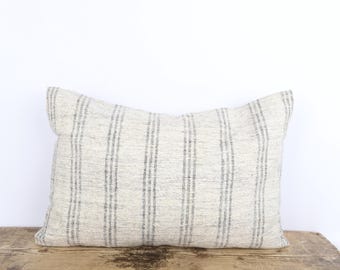 May include: Rectangular pillow with a cream-colored background and vertical gray stripes. The pillow is made of a textured fabric and rests on a wooden surface. A decorative accent for a bed or sofa.