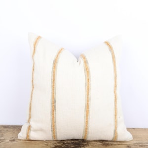 Peut inclure: Coussin carré crème avec des rayures verticales beiges et grises. Le coussin est posé sur une surface en bois rustique sur fond blanc. La texture et le design du coussin suggèrent un élément de décoration intérieure confortable.
