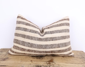 May include: A rectangular pillow with horizontal stripes in alternating shades of brown and cream. The pillow is made of a textured fabric and rests on a wooden surface. The pillow is a decorative home accent.