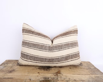May include: A rectangular pillow with a cream-colored background and horizontal brown stripes. The pillow is placed on a weathered wooden surface against a white backdrop. The pillow's design is simple and rustic.