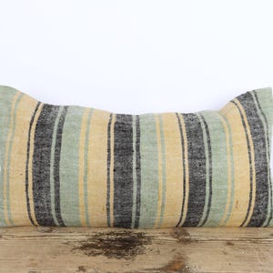 Peut inclure: Coussin rectangulaire décoratif à rayures. Le coussin présente des rayures verticales dans des tons de jaune, vert et noir. Il est posé sur une surface en bois sur fond blanc.