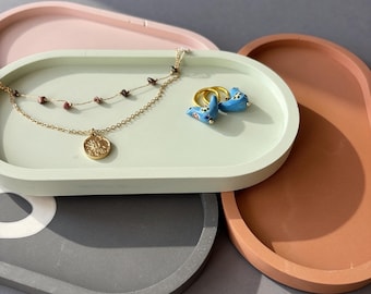 May include: A collection of four oval-shaped trays in pastel pink, light green, terracotta, and dark gray. The light green tray holds a gold-toned chain necklace with small dark beads and a round pendant, alongside two blue ceramic hoop earrings.