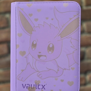Op de afbeelding: Een lichtpaarse kaartenmap met een lijntekening van het personage Eevee omringd door kleine hartjes. De kaft heeft een gestructureerde afwerking en de merknaam vaultx staat in donkerpaarse letters in de linkerbenedenhoek gedrukt.