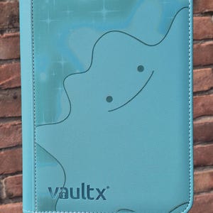Può includere: Un raccoglitore per carte azzurro con superficie testurizzata che presenta il contorno in rilievo di una creatura simile a una macchia con due occhi circolari e una bocca curva. La parola vaultx è stampata nell'angolo in basso a sinistra.