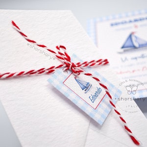 May include: A white envelope with a red and white twine bow. A blue and white gift tag with a sailboat and the name "Edoardo" is attached to the bow. The tag has a blue and white gingham pattern.