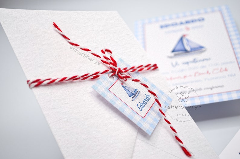 May include: A white envelope with a red and white twine bow. A blue and white gift tag with a sailboat and the name "Edoardo" is attached to the bow. The tag has a blue and white gingham pattern.