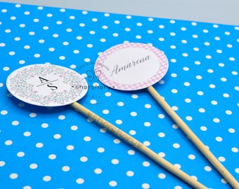 以下が含まれることがあります： 木製の棒が付いた丸いケーキトッパーが2つ。1つは白い花輪と「A/S」のイニシャルが入った白いトッパー。もう1つは、白いギンガムチェックの縁取りと、筆記体で「Amarena」と書かれたピンクのトッパーです。