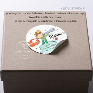 May include: A beige gift box with a round sticker featuring a cartoon illustration of a boy with a telescope, a red scarf, and a small rose. The sticker says "Mattia" and "Prima Comunione 14.03.2022".