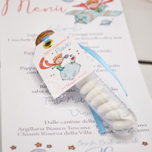 Puede incluir: Un peque&ntilde;o tubo de pl&aacute;stico transparente lleno de caramelos blancos. El tubo est&aacute; atado a una tarjeta blanca con una cinta azul. La tarjeta presenta una ilustraci&oacute;n de dibujos animados de un ni&ntilde;o con una capa roja y un sombrero azul, de pie en un planeta con una flor roja. El texto en la tarjeta dice "Santo Battesimo di Flavio 11 Maggio 2022".