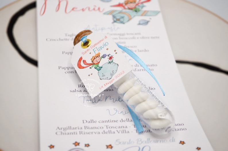 Puede incluir: Un peque&ntilde;o tubo de pl&aacute;stico transparente lleno de caramelos blancos. El tubo est&aacute; atado a una tarjeta blanca con una cinta azul. La tarjeta presenta una ilustraci&oacute;n de dibujos animados de un ni&ntilde;o con una capa roja y un sombrero azul, de pie en un planeta con una flor roja. El texto en la tarjeta dice "Santo Battesimo di Flavio 11 Maggio 2022".