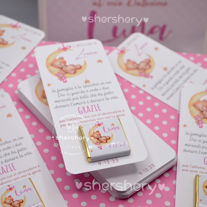 May include: A white card with a pink and white polka dot background. The card features a cartoon illustration of a teddy bear sleeping on a crescent moon. The card has the text "GRAZIE" and "Luna" in gold lettering. The card is accompanied by a small chocolate bar with a gold wrapper and the text "Luna" and "9.12.23" printed on it.