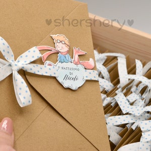 May include: A brown envelope with a white ribbon tied in a bow. The ribbon has blue polka dots. The envelope has a sticker with an illustration of a boy in a blue jacket and a red scarf riding a fox. The sticker also has the text "Battesimo di Nicol&ograve;".