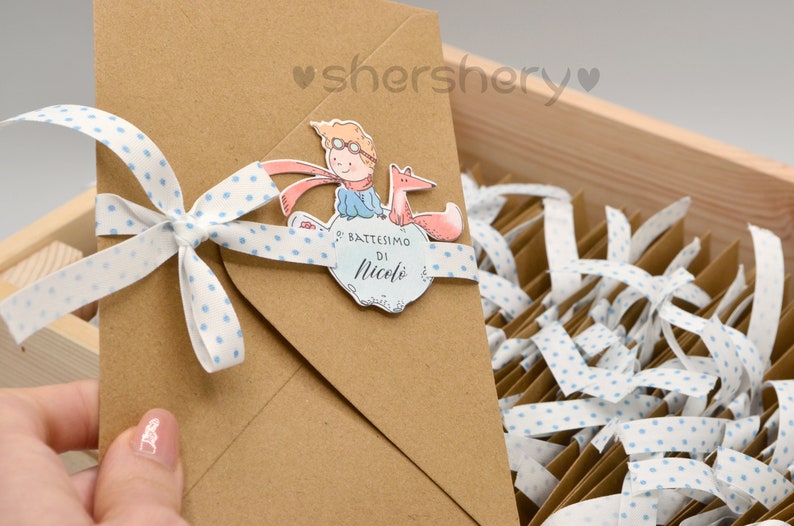 May include: A brown envelope with a white ribbon tied in a bow. The ribbon has blue polka dots. The envelope has a sticker with an illustration of a boy in a blue jacket and a red scarf riding a fox. The sticker also has the text "Battesimo di Nicol&ograve;".