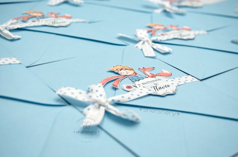 May include: A stack of light blue envelopes with white polka dot ribbon and a small illustration of a boy with a fox. The envelope has the text "Il Battesimo di  [Name]" printed on it.