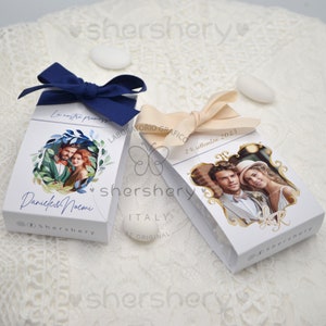 Puede incluir: Dos cajas blancas para recuerdos con fotos de una pareja, atadas con cintas azul marino y crema. Una caja tiene una corona de acuarela y los nombres "Danieles & Noemi". La otra caja tiene la fecha "24 settembre 2023."
