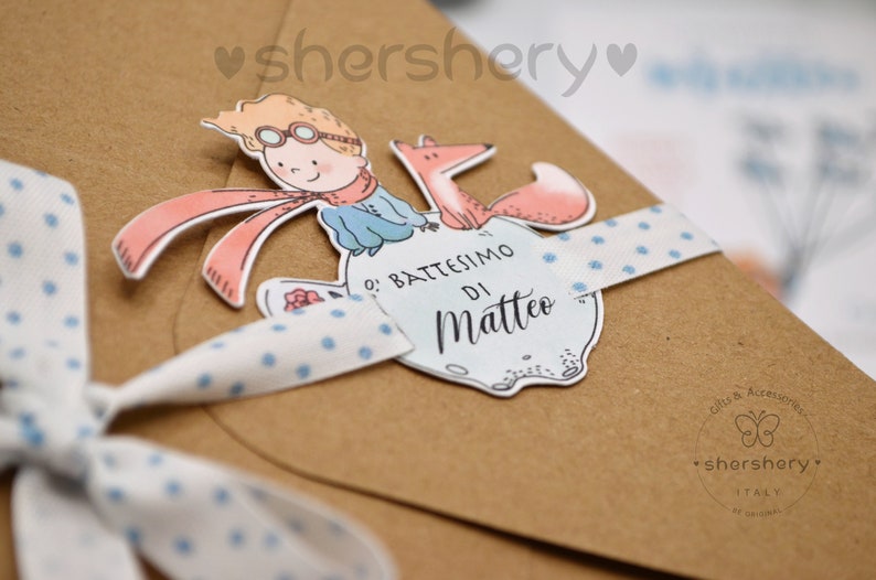 May include: A brown paper envelope with a blue and white polka dot ribbon tied around it. The envelope has a sticker on it with a cartoon image of a boy with a fox. The sticker says "O' Battesimo di Matteo".