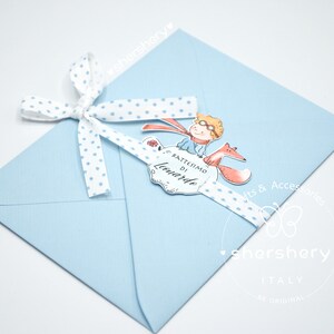 May include: A light blue envelope with a white ribbon tied around it. The ribbon has blue polka dots. A small card with a cartoon illustration of a boy and a fox is attached to the ribbon. The card says "Battesimo di Leonardo".