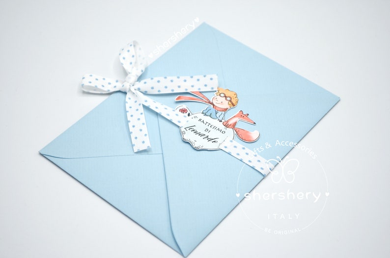 May include: A light blue envelope with a white ribbon tied around it. The ribbon has blue polka dots. A small card with a cartoon illustration of a boy and a fox is attached to the ribbon. The card says "Battesimo di Leonardo".