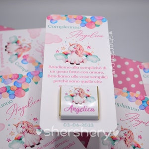 May include: A white card with a unicorn illustration and the text "Compleanno Angelica" and "Brindiamo alla semplicit&agrave; di un gesto fatto con amore, Brindiamo alle cose semplici perch&egrave; sono quelle che". The card also has a small chocolate bar with a unicorn illustration and the text "Angelica" and the date "03-06-2023".