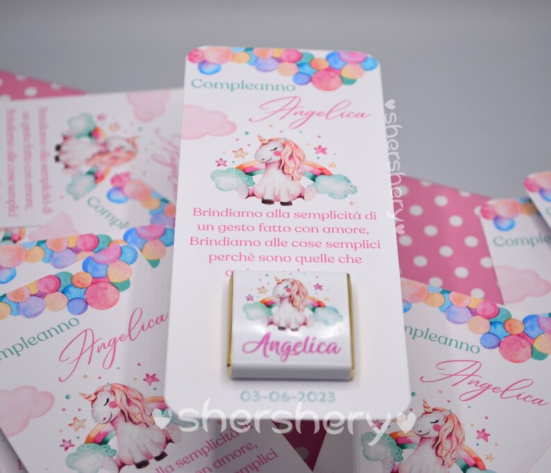 May include: A white card with a unicorn illustration and the text "Compleanno Angelica" and "Brindiamo alla semplicit&agrave; di un gesto fatto con amore, Brindiamo alle cose semplici perch&egrave; sono quelle che". The card also has a small chocolate bar with a unicorn illustration and the text "Angelica" and the date "03-06-2023".