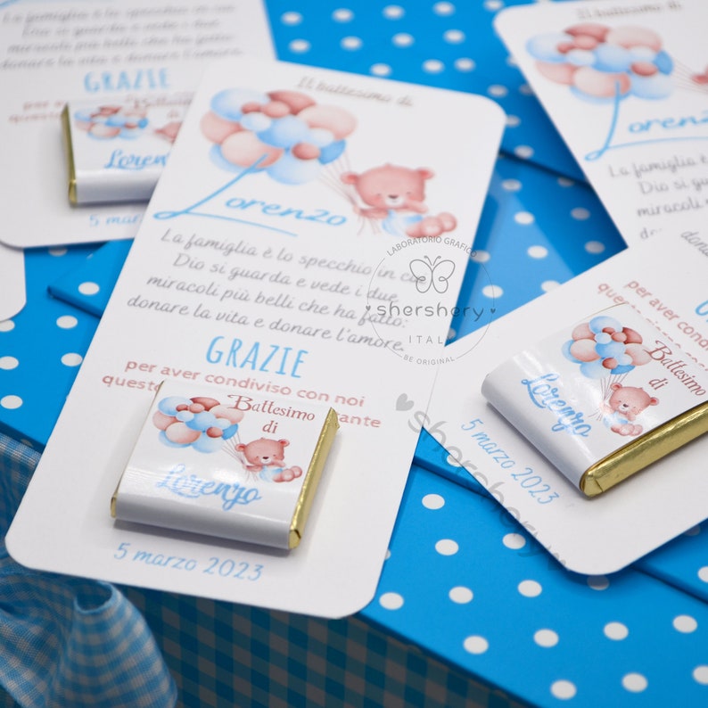 May include: A white card with blue polka dots and a blue and white checkered pattern on the back. The card has a white chocolate bar with a picture of a teddy bear holding balloons. The text on the card reads "GRAZIE" and "Battesimo di Lorenzo" with the date "5 marzo 2023".