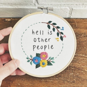 Peut inclure: Art de broderie sur un cercle avec la citation "l'enfer, c'est les autres" entourée de feuilles vertes et de fleurs colorées.