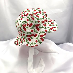 May include: A white sun hat with a cherry print. The hat has a wide brim and a tie that can be tied under the chin.