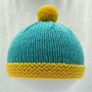 May include: A teal blue knitted hat with a yellow pom pom on top and a yellow brim.