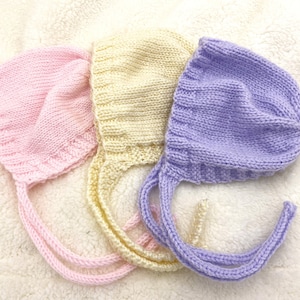 May include: Three knitted baby bonnets in pink, yellow, and purple with ties. The bonnets are made of soft yarn and have a classic style.