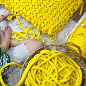 May include: A large yellow knitted blanket made with thick yarn. The yarn is also shown in a basket and loose on the floor.