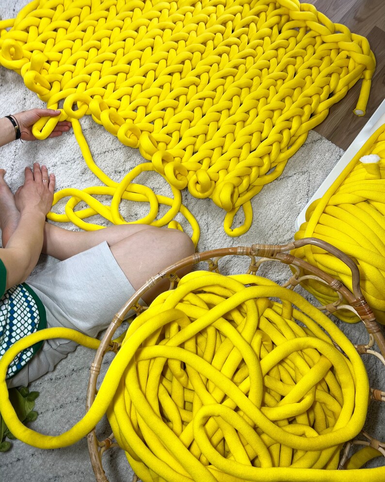 May include: A large yellow knitted blanket made with thick yarn. The yarn is also shown in a basket and loose on the floor.