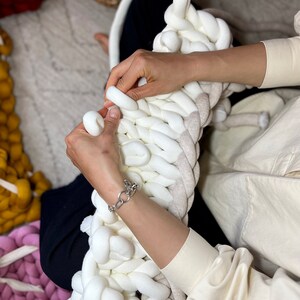 May include: A person is knitting a large, white and beige chunky knit blanket. The blanket is made with thick yarn and has a soft, fluffy texture.