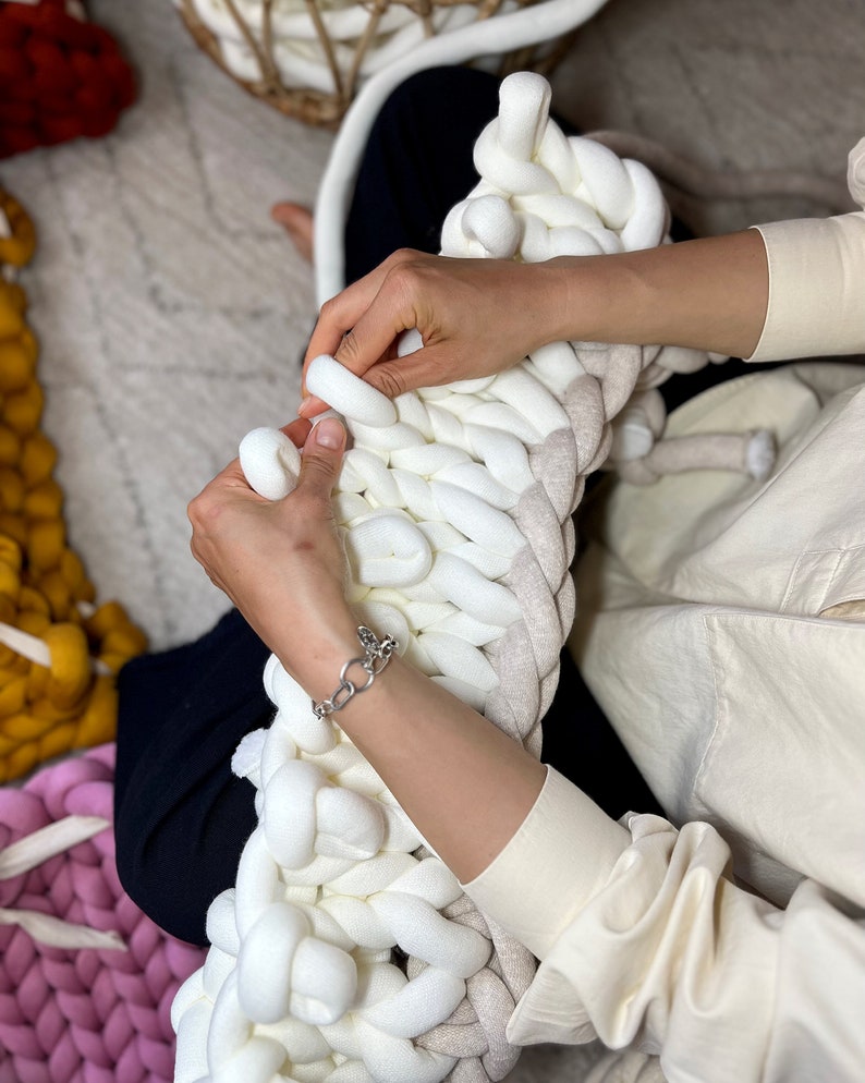 May include: A person is knitting a large, white and beige chunky knit blanket. The blanket is made with thick yarn and has a soft, fluffy texture.