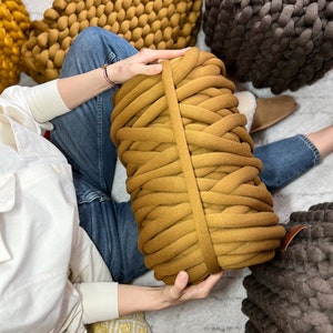 May include: A large roll of mustard yellow chunky yarn with a brown leather strap. The yarn is sitting on a white surface with a person's hand holding it.