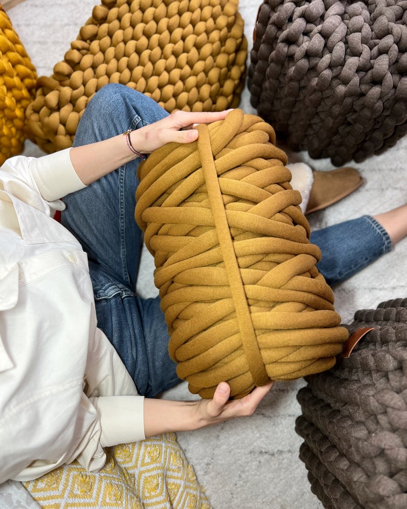 May include: A large roll of mustard yellow chunky yarn with a brown leather strap. The yarn is sitting on a white surface with a person's hand holding it.