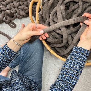 May include: A person is holding a large ball of brown yarn in a wicker basket. The yarn is thick and chunky, and the person is holding it in their hands, ready to start knitting or crocheting.