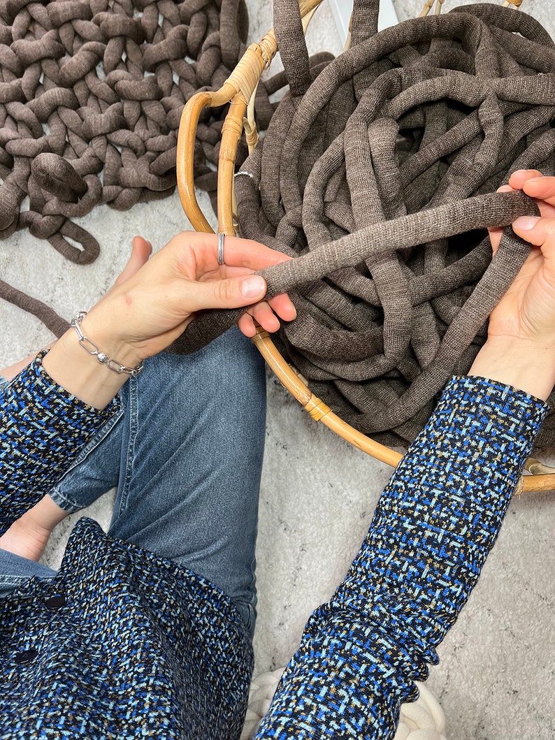 May include: A person is holding a large ball of brown yarn in a wicker basket. The yarn is thick and chunky, and the person is holding it in their hands, ready to start knitting or crocheting.