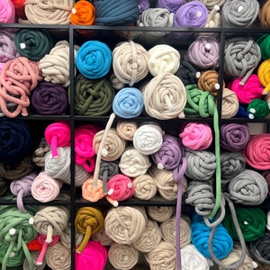 May include: A variety of colorful yarn balls are displayed on shelves. The yarn is made of thick, chunky material and comes in a range of colors, including pink, blue, green, gray, and white.