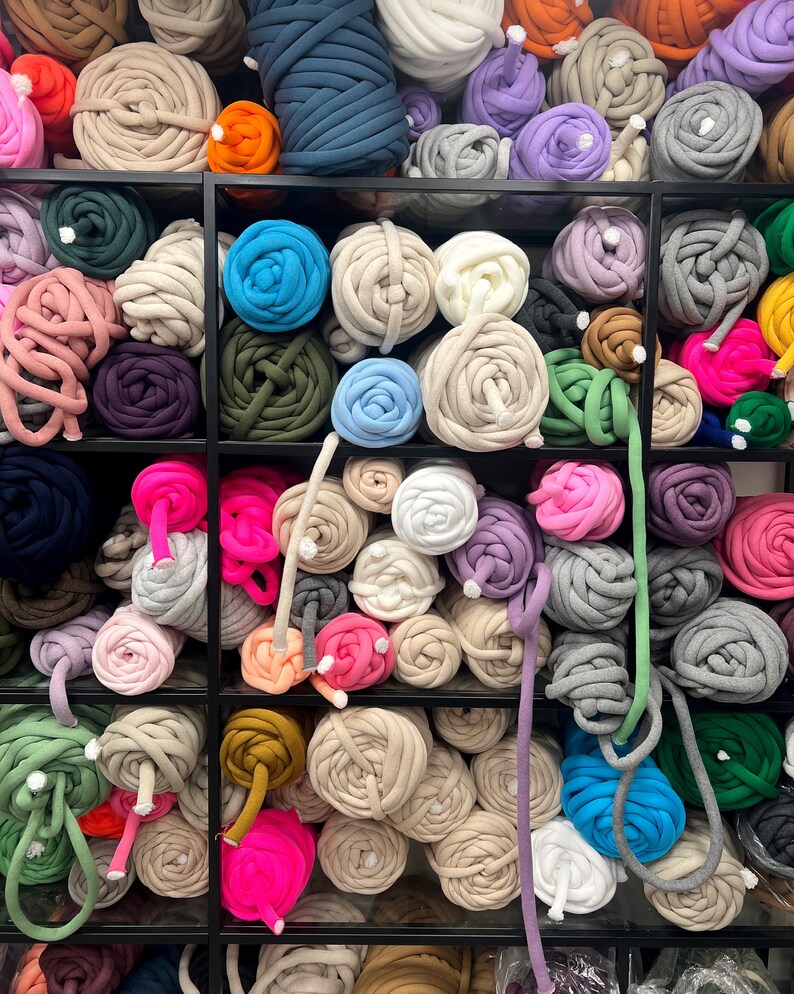 May include: A variety of colorful yarn balls are displayed on shelves. The yarn is made of thick, chunky material and comes in a range of colors, including pink, blue, green, gray, and white.
